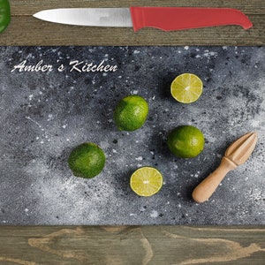 Peut inclure: Une planche à découper noire avec une finition mouchetée blanche. La planche porte l'inscription "Amber's Kitchen". Il y a cinq citrons verts sur la planche, quatre entiers et un coupé en deux. Un presse-citron en bois se trouve également sur la planche.