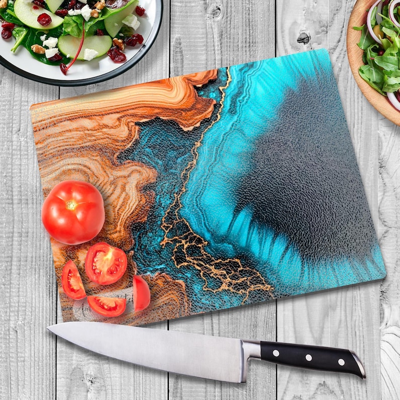 May include: A blue and brown abstract design glass cutting board with gold accents. The board is on a wooden surface with tomatoes and a knife.