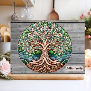 Può includere: Un pannello decorativo in legno con un colorato disegno dell'albero della vita. L'opera d'arte presenta un albero dettagliato con rami, foglie e radici intricati, su uno sfondo di venature del legno grigio. Il pannello ha il testo "Saltos Family".