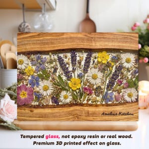 Puede incluir: Tabla de cortar rectangular con marco de madera y diseño floral. El centro presenta una variedad de flores prensadas en tonos amarillos, morados, blancos y rosas. El texto "Amelias Kitchen" es visible en la parte inferior derecha.