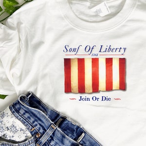 May include: White t-shirt with a red, white, and blue striped flag design. The text "Sons of Liberty 1765" is above the flag and "Join or Die" is below the flag.