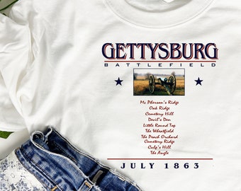 May include: White t-shirt with a blue and red graphic design that says "Gettysburg Battlefield" with a picture of a cannon and the text "July 1863".