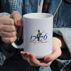 Puede incluir: Taza de cerámica blanca con un gráfico azul y rojo de una persona sosteniendo un rifle con el texto "1776 Liberty & Freedom".