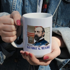 May include: White ceramic coffee mug with a portrait of Major General George G. Meade, a Union general in the American Civil War. The mug features a red, white, and blue American flag and text that reads "MAJOR GENERAL GEORGE G. MEADE".