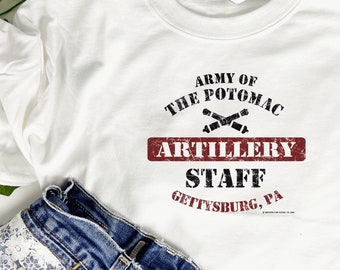 May include: White t-shirt with the text "ARMY OF THE POTOMAC ARTILLERY STAFF GETTYSBURG, PA" in black and red. The shirt is paired with blue denim jeans with embroidered hearts and lace detailing.