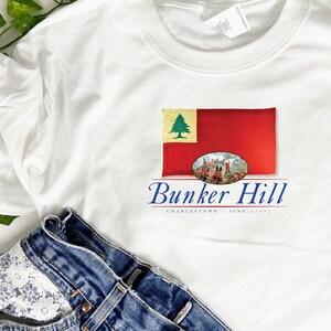 May include: White t-shirt with a red, white, and green flag design and the text "Bunker Hill, Charlestown - June 17, 1775".