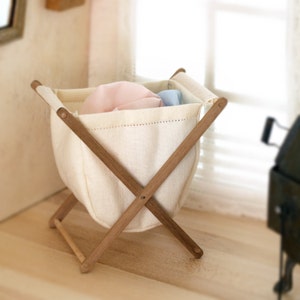 Op de afbeelding: Een witte canvas wasmand met een houten frame. De mand is gevouwen en heeft een roze en blauwe stof erin.