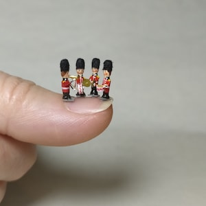 May include: A miniature toy band of four figures in red jackets, black hats, and white trousers. One figure holds a trumpet, another a cymbal, and a third a drum. The figures are standing on a finger.