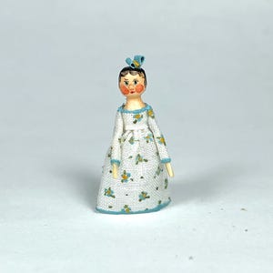 May include: A miniature doll with a painted face, wearing a white dress with blue floral patterns and trim. The doll has a blue bow in her hair and wooden arms. The doll is a charming decorative item.