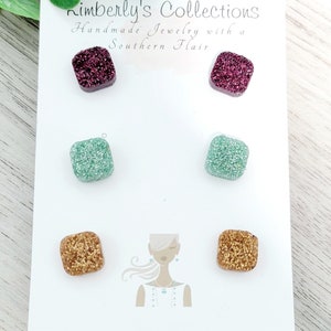 May include: A set of six square-shaped stud earrings with glitter embedded in the resin. The earrings are in three colors: burgundy, teal, and gold. The earrings are displayed on a white card with the text "Kimberly's Collections Handmade Jewelry with a Southern Flair".