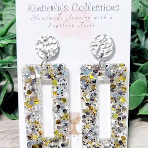 May include: Silver and gold glitter rectangle earrings with a silver textured disc charm. The earrings are handmade and have a Southern Flair. Kimberly's Collections.