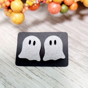 May include: Two silver glitter ghost earrings on a black background. The ghosts have two small black eyes.