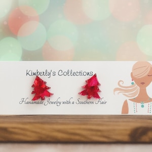 May include: A pair of red Christmas tree earrings displayed on a white card with the text "Kimberly's Collections Handmade Jewelry with a Southern Hair". The background is a blurred bokeh of green, red, and white lights.