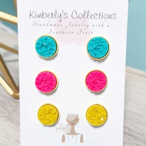 May include: Three pairs of stud earrings on a white card with the text "Kimberly's Collections Handmade Jewelry with a Southern Flair". The earrings are turquoise, pink, and yellow.