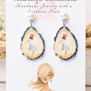 May include: A pair of oyster shell earrings featuring a painted stork wearing a red, white, and blue top hat. The earrings are attached to a white display card printed with the text Kimberly's Collections Handmade Jewelry with a Southern Flair.