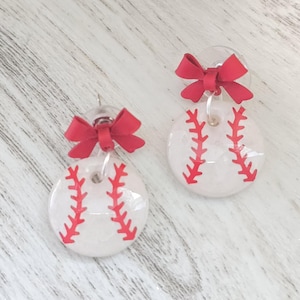 May include: A pair of baseball-themed earrings. Each earring features a white, circular design with red stitching, resembling a baseball. A small, red bow is attached to the top of each earring.