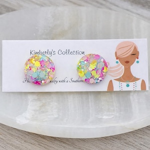 May include: A pair of round stud earrings with colorful glitter embedded in clear resin. The earrings are displayed on a white card with the text "Kimberly's Collection" and a graphic of a woman.