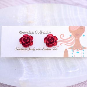 May include: A pair of red rose stud earrings on a white card. The card reads "Kimberly's Collection" and "Handmade Jewelry with a Southern Flair." The earrings are on a white surface.
