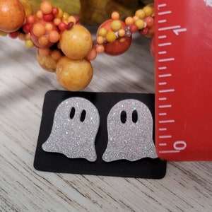 May include: Two white glitter ghost earrings on a black card with a ruler for size reference. The ghosts are approximately 1 inch tall.