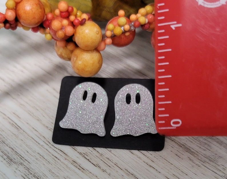 May include: Two white glitter ghost earrings on a black card with a ruler for size reference. The ghosts are approximately 1 inch tall.
