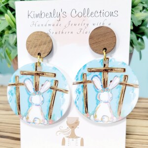 May include: A pair of wooden and painted earrings featuring a white bunny rabbit in front of three wooden crosses. The earrings are blue, white, and brown. The background is a light blue sky with white clouds. The earrings are on a white card with the text "Kimberly's Collections Handmade Jewelry with a Southern Flair".