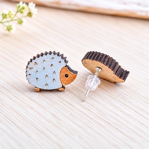 May include: Wooden hedgehog stud earrings. The earrings feature a light blue body with brown accents and a dark brown spiky back. The earrings have a post and a clear earring back. The earrings are on a light wood surface.