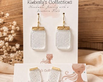 May include: Two pairs of earrings on display cards labeled Kimberly's Collections. Each earring features a clear, rectangular mason jar shape filled with silver glitter, topped with a textured gold-colored metal cap and a gold-colored hook wire.