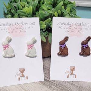 May include: Two pairs of bunny rabbit stud earrings. One pair is white with pink bows and the other pair is brown with purple bows. The earrings are displayed on white cards with the text "Kimberly's Collections Handmade Jewelry with a Southern Flair".