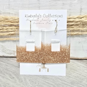 May include: A pair of gold glitter rectangle earrings with a clear acrylic frame and silver hooks. The earrings are displayed on a white card with the text "Kimberly's Collections Handmade Jewelry with a Southern Flair".