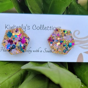 May include: A pair of hexagonal stud earrings with a glittery design. The earrings feature gold, pink, teal, and purple glitter. The earrings are displayed on a white card with the text "Kimberly's Collections".