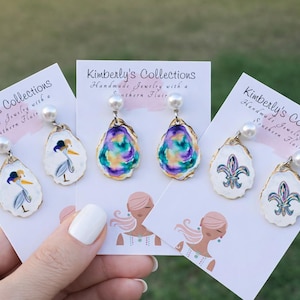 May include: Handmade oyster shell earrings with pearl accents. Designs include a crane, a fleur-de-lis, and abstract colorful patterns. Each earring is attached to a white card with the text "Kimberly's Collections Handmade Jewelry with a Southern Flair."