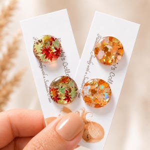 May include: Four circular resin stud earrings displayed on two white cards. Two earrings feature red, green, and gold leaf-shaped glitter, while the other two contain orange, gold, and iridescent hexagonal glitter. The cards include text.