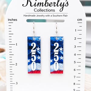 May include: Rectangular earrings with a patriotic design. The earrings feature a blue, white, and red color scheme with the number "250" in white. The earrings are approximately 2 inches long.