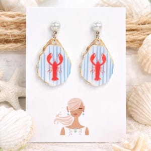 May include: Lobster-themed earrings. Each earring features a white and blue striped background with a red lobster design. The earrings are made from oyster shells and have a pearl accent at the top. The earrings are displayed on a white card with a woman's illustration.