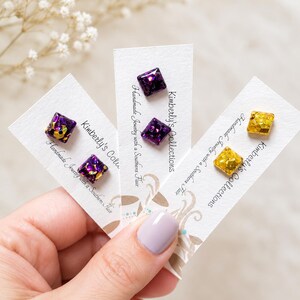 May include: Three pairs of square stud earrings displayed on white cards labeled Kimberly's Collections. The earrings feature purple and gold glitter resin designs. Each card is printed with the text Handmade Jewelry with a Southern Flair.