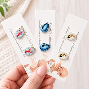 May include: Three pairs of handmade earrings on white cards. One pair features a red lobster design on a blue and white striped background. The other two pairs have blue oyster and white oyster designs. The cards read "Kimberly's Collections."
