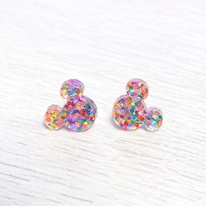 May include: Two glittery, colorful Mickey Mouse-shaped stud earrings. The earrings are filled with a variety of small, colorful glitter pieces, creating a sparkling effect. The earrings are set against a white background.