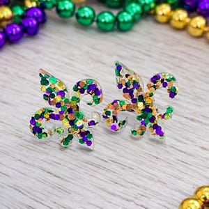 May include: Clear resin fleur-de-lis earrings filled with green, purple, and gold glitter. The earrings are on a light wood surface, with Mardi Gras beads in the background.