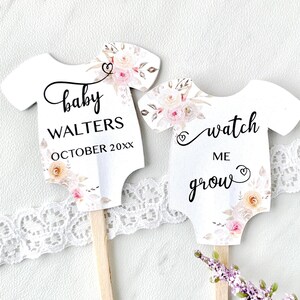 May include: Two white baby onesie shaped cupcake toppers with floral designs. The first topper says "baby WALTERS OCTOBER 20XX" and the second topper says "watch me grow".