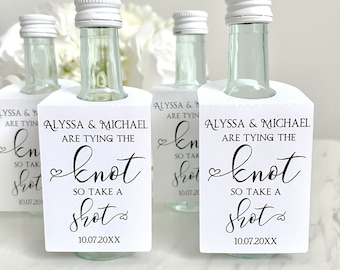 May include: Clear glass mini liquor bottles with white screw caps, each featuring a white rectangular paper tag printed with black text that reads: Alyssa & Michael are tying the knot so take a shot, including the date 10.07.20XX.
