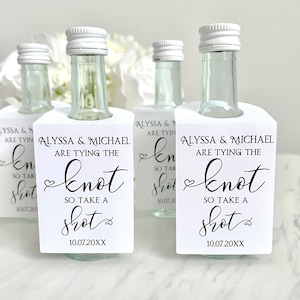 May include: Four small glass bottles with white caps and white labels. The labels have black text that reads "Alyssa & Michael are tying the knot so take a shot" and the date "10.07.20XX".
