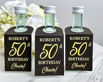 May include: Three small glass bottles with silver caps, each in a black holder. The holders have gold text that reads "ROBERT'S 50th BIRTHDAY Cheers!". The bottles are on a white marble surface.