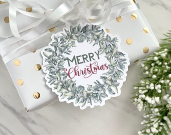 May include: A white gift box with gold polka dots and a white ribbon. A round gift tag with a green wreath and the text "Merry Christmas" in red lettering is attached to the box.