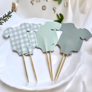 May include: Four green and white baby onesie cupcake toppers with wooden sticks. The onesies are in different patterns, including gingham and solid.