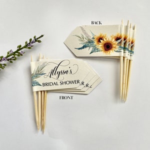 May include: Set of six bridal shower cake toppers with a sunflower and greenery design. The front of the topper says "Alyssa's Bridal Shower". The back of the topper is blank.