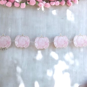 May include: A garland of pink pumpkin decorations, each about 11.4 cm wide, strung on a white thread. The pumpkins feature a glitter design and are set against a light grey backdrop, with pink pom-poms at the top, creating a festive look.