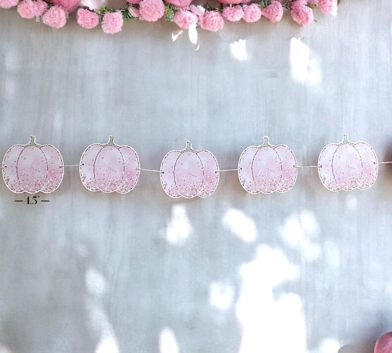 May include: A garland of pink pumpkin decorations, each about 11.4 cm wide, strung on a white thread. The pumpkins feature a glitter design and are set against a light grey backdrop, with pink pom-poms at the top, creating a festive look.