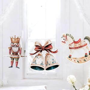 May include: A festive garland featuring watercolor cutouts of a nutcracker, bells, and a rocking horse. The nutcracker is in a red and gold uniform, the bells have a bow, and the rocking horse has a red saddle. The garland is strung on a white string.