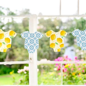 May include: A garland of baby onesie cutouts hangs in front of a window. Two onesies feature yellow lemons on a yellow background, while the other two have a blue and white patterned design. The background shows a blurred view of greenery and flowers.