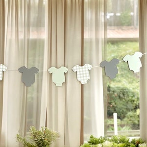 Op de afbeelding: Een baby shower banner met afwisselend grijze en groene baby rompers. De rompers zijn uitgesneden en aan elkaar geregen met wit touw.